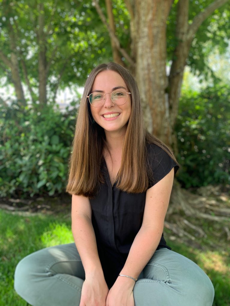 Taylor Nonnenmacher, RDN Young woman with glasses smiling, seated outdoors in front of a tree and greenery.