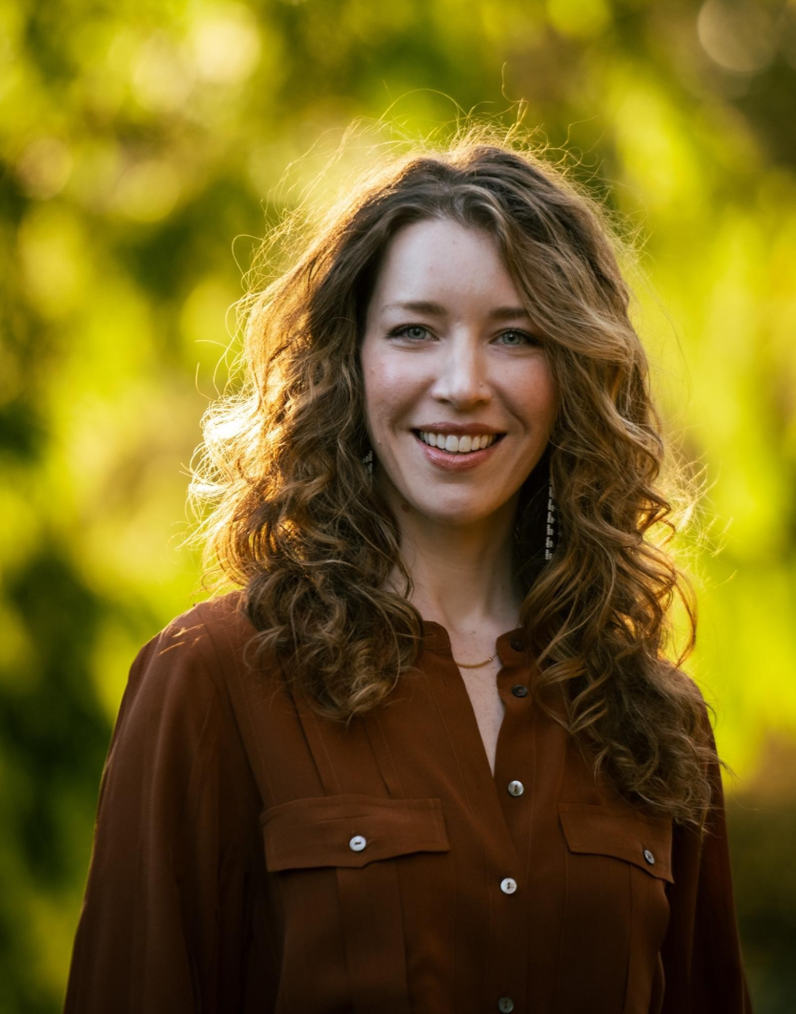 Julia Hollenberg A woman with long curly hair and wearing a rust-colored blouse stands outdoors with greenery in the background, smiling at the camera.