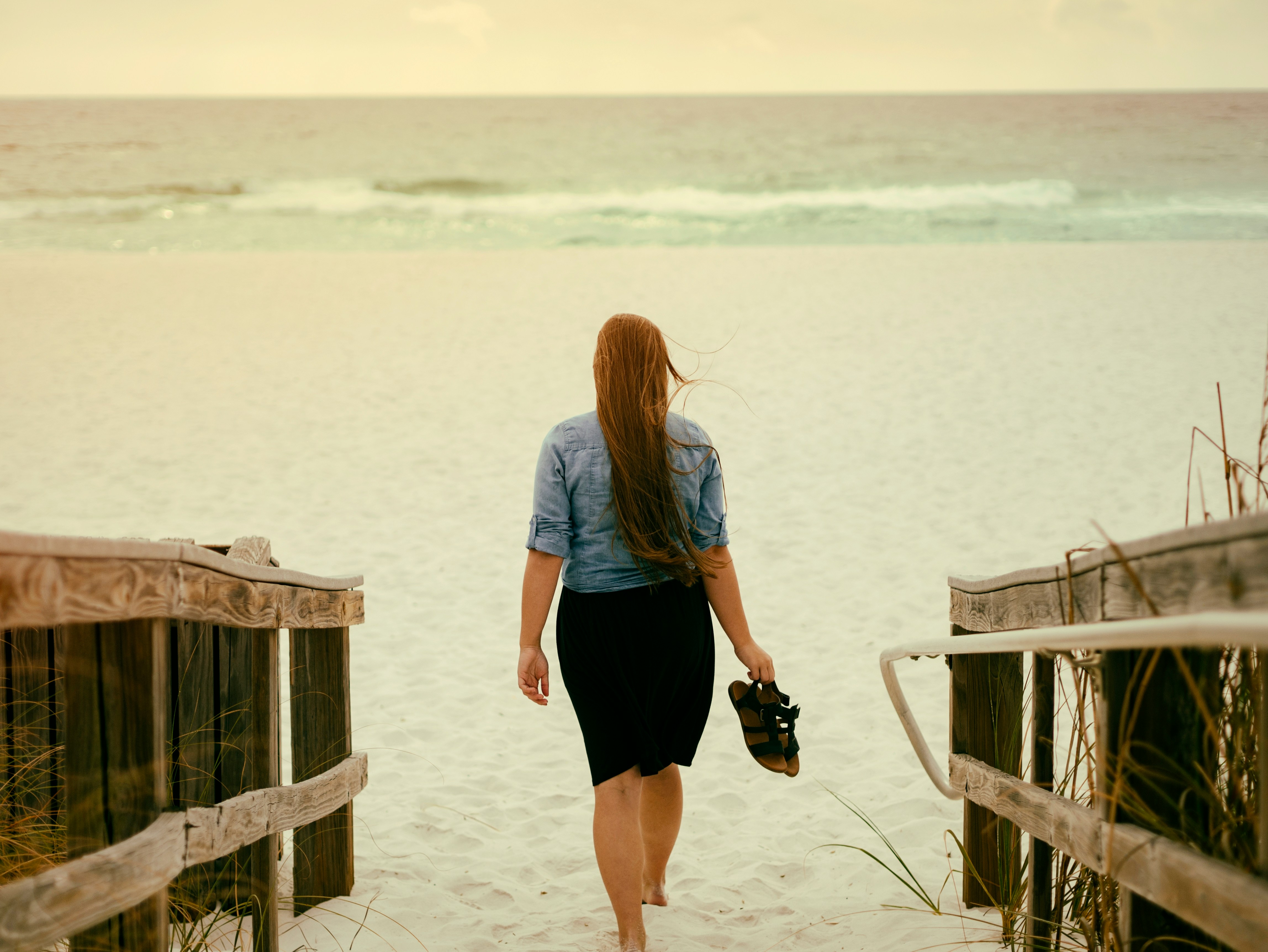aaron-burden-z9Y7GXQ37lg-unsplash A person with long hair, wearing a light blue shirt and dark skirt, walks down a wooden pathway towards a sandy beach, holding shoes in one hand. The ocean is visible in the background under a slightly overcast sky.